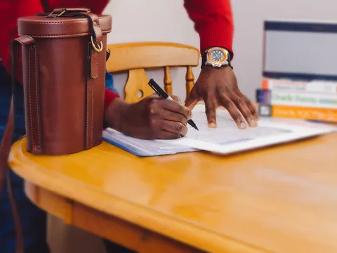 Hand, die mit einem Stift ein ausgedrucktes Formular auf einem Holztisch ausfüllt